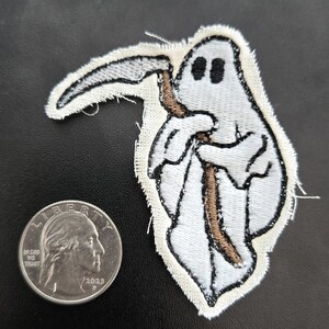 May include: An embroidered patch featuring a white ghost holding a brown scythe with a silver blade. The patch is shown next to a 2023 United States quarter coin for scale, highlighting the size of the ghost figure against the standard currency.