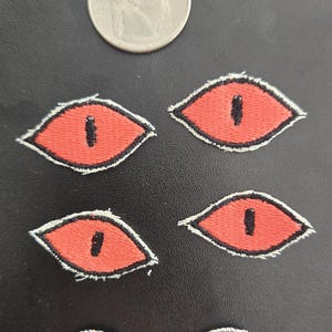 May include: Six embroidered patches shaped like reptilian eyes with coral-red irises and vertical black pupils, outlined in white and black thread. A silver-colored United States quarter coin is positioned above the patches for scale reference.