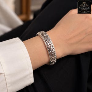 Pode incluir: Uma pulseira rígida de tom prateado com um intrincado padrão de filigrana vazada com recortes em espiral. A superfície metálica tem um acabamento polido. O logótipo Avora Jewels é visível no canto superior direito da imagem.