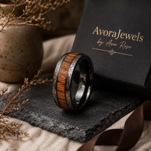 May include: A ring featuring a central band of brown wood grain inlay flanked by dark metal edges with intricate silver-toned floral engraving. The ring is positioned near a black box with the gold text AvoraJewels by Ava Rose printed on it.
