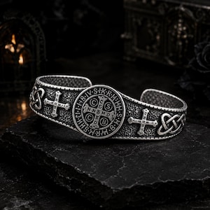 Puede incluir: Un brazalete de metal plateado con una medalla central circular de San Benito con letras y una cruz en relieve. La banda tiene un acabado texturizado y está decorada con símbolos de cruces y patrones de nudos celtas repetidos.