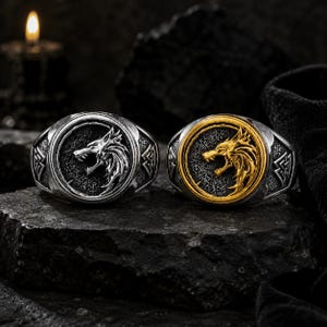 May include: Two rings featuring a circular face with a relief of a snarling wolf head. One ring is silver-toned, and the other is gold-toned with a silver-toned band. Both rings have a textured dark background and geometric side engravings.