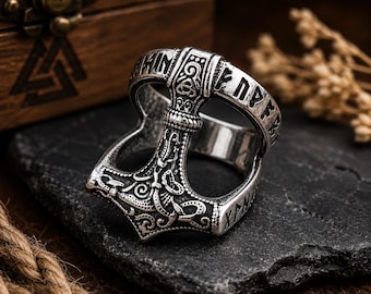 Puede incluir: Un anillo de metal plateado con forma de martillo de Mjolnir, que presenta intrincados patrones de nudos en relieve y caracteres rúnicos grabados a lo largo de la banda. El diseño incorpora un motivo central de cabeza de martillo con volutas detalladas y acentos oxidados.