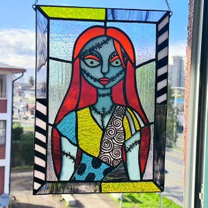May include: A rectangular stained glass panel featuring a portrait of a character with long red hair, pale blue skin, and stitched facial details. The figure wears a patchwork dress with yellow, blue, and spiral patterns, framed by black and white borders.
