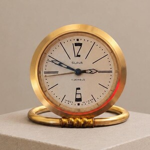 May include: A round gold-tone analog desk clock with a cream-colored face. The dial features black numerals at the 12 and 6 positions, black hour markers, and black hands. The text SLAVA, 11 JEWELS, and MADE IN USSR is printed on the clock face.