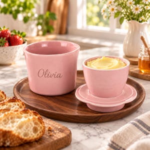 May include: A pink ceramic butter keeper set consisting of a base and a lid, with the name Olivia engraved on the side of the base. The lid holds a portion of yellow butter, and both pieces are arranged on a round wooden serving tray.