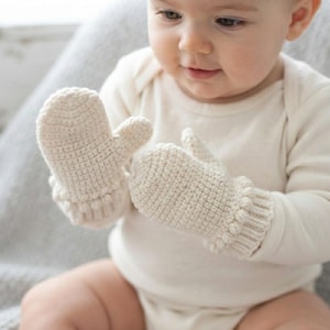 May include: A pair of cream-colored crocheted baby mittens featuring a textured stitch pattern and ribbed cuffs. The mittens are crafted from a soft yarn and are designed to cover the hands, providing a simple and functional accessory for infants.