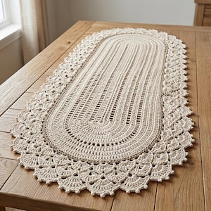 May include: An oval-shaped, cream-colored crochet table runner featuring a central textured pattern with vertical lines and a wide, decorative scalloped border with intricate shell-like stitching along the entire outer edge of the piece.