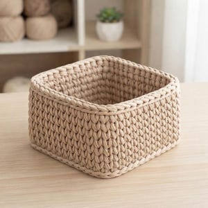 May include: A square-shaped storage basket crafted from thick, light beige yarn in a chunky crochet knit pattern. The basket features a sturdy, open-top design with a consistent, textured weave throughout the sides and a reinforced rim edge.