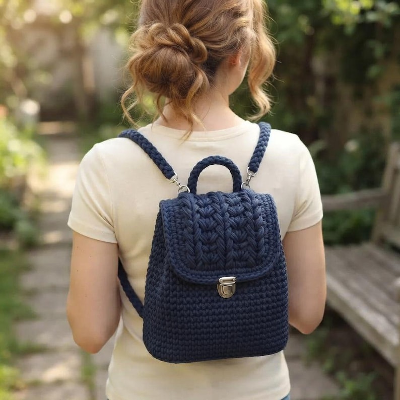 May include: A dark navy blue crochet backpack featuring a textured braided pattern on the top flap, a sturdy top handle, and a silver-toned metal clasp closure. The bag has thick, braided shoulder straps attached with silver-toned metal hardware.