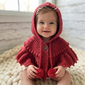 May include: A deep red crochet hooded capelet featuring a textured bobble stitch pattern, a single wooden button closure at the neck, and two matching red yarn pom-poms attached to the ends of the hood drawstrings, creating a cozy garment.