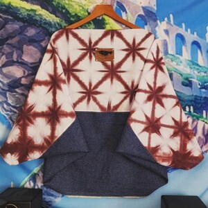 May include: A wide-cut top featuring a cream-colored upper section with a dark red starburst tie-dye pattern and a solid dark blue lower section. A small rectangular brown leather patch with a black graphic is centered on the upper back.