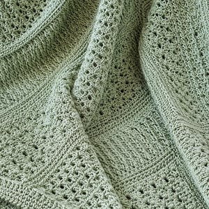 May include: A sage green crocheted blanket featuring a complex pattern of alternating solid stitches and openwork diamond-shaped eyelets. The textile displays a textured surface with visible yarn loops and a consistent, uniform sage green color throughout.