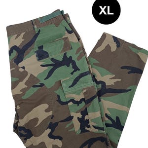 Op de afbeelding: Cargobroek met een woodland camouflagepatroon in tinten groen, bruin en zwart. De broek heeft een zijzak met klepsluiting en een tailleband met riemlussen. Een zwart cirkelvormig icoon met de witte tekst XL is zichtbaar in de afbeelding.