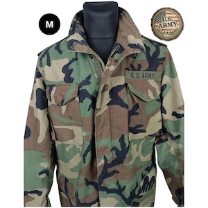 May include: A camouflage field jacket featuring a woodland pattern of green, brown, and black. The jacket includes a high collar, a front zipper closure, and two chest pockets with flaps. A patch reading U.S. ARMY is above the left pocket.
