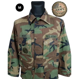 Könnte beinhalten: Eine langärmelige Tarnjacke mit Waldmuster in Grün-, Braun- und Schwarztönen. Die Jacke verfügt über zwei Brusttaschen mit Klappen, einen Kragen und zwei gestickte Aufnäher mit der Aufschrift MORE und U.S. ARMY. Ein Etikett mit Größe M ist sichtbar.