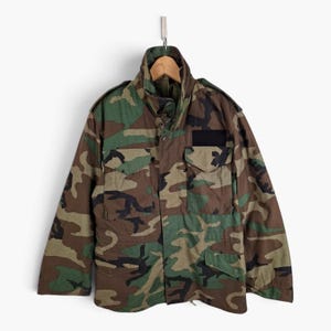 May include: A field jacket featuring a woodland camouflage pattern in shades of green, brown, and black. The jacket includes a high collar, a front zipper closure with a buttoned storm flap, two chest pockets with flaps, and a black rectangular patch.