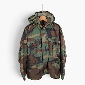 May include: A woodland camouflage field jacket featuring a hood, a front zipper closure with a buttoned storm flap, and four large front pockets. The jacket includes a patch on the upper left sleeve and long sleeves with adjustable cuffs.