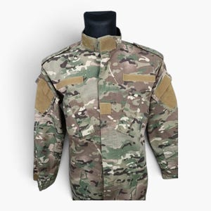 Könnte beinhalten: Eine langärmelige taktische Jacke mit einem mehrfarbigen Tarnmuster in Grün-, Braun- und Beigetönen. Die Jacke verfügt über einen hohen Kragen mit Klettverschluss, mehrere Brusttaschen sowie beigefarbene Klettflächen an Brust und Ärmeln.