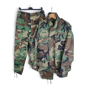 May include: A camouflage field jacket and matching cargo trousers featuring a woodland pattern of green, brown, and black. The jacket has a high collar, front pockets, and the text U.S. ARMY embroidered in black on the chest area.