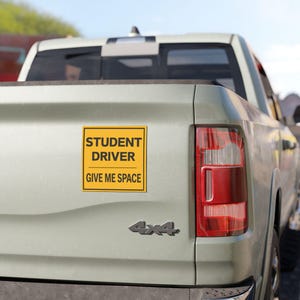 Op de afbeelding: Een vierkante gele sticker met een zwarte rand op de achterklep van een lichtgekleurde pick-up truck. De sticker bevat de zwarte tekst "STUDENT DRIVER" boven een horizontale lijn, met de tekst "GIVE ME SPACE" eronder gedrukt.