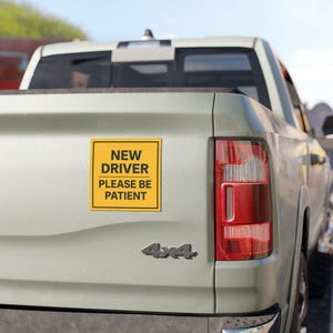 Peut inclure: Un autocollant carré jaune avec une fine bordure noire apposé sur le hayon d'un pick-up de couleur claire. L'autocollant présente un texte noir en gras qui indique NEW DRIVER PLEASE BE PATIENT, séparé par une ligne horizontale entre les phrases.
