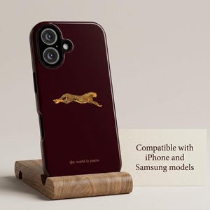 May include: A dark burgundy smartphone case featuring a small, centered graphic of a running cheetah in gold and black tones. Below the cheetah, the text reads "the world is yours" in small gold letters. The case is compatible with iPhone and Samsung models.