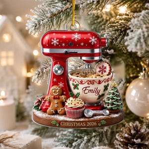 May include: A red stand mixer Christmas ornament featuring white snowflake patterns, a gingerbread person, a cupcake, and a mixing bowl with the name Emily printed on it. The base is wood-toned with the text CHRISTMAS 2026 and a small tree.