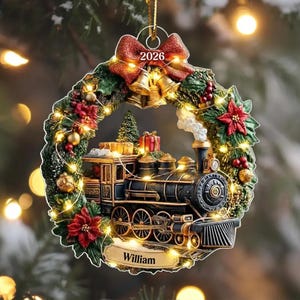 May include: A circular Christmas ornament featuring a steam locomotive train inside a wreath of green pine needles, red poinsettias, and gold bells. A red bow at the top displays the year 2026, and a gold plaque at the bottom is inscribed with the name William.