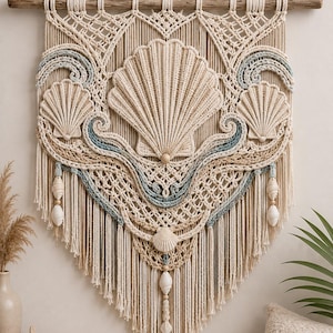 May include: A macrame wall hanging suspended from a wooden branch, featuring intricate cream-colored cord knots, blue wave-like accents, and three large central shell motifs. The design includes hanging shell charms and a long, fringed bottom edge.