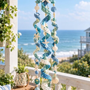 May include: A hanging spiral mobile featuring three crocheted yarn strands in shades of blue, teal, and white. The strands are adorned with small wooden beads, white starfish, and various seashells, suspended from a circular wooden ring at the top.