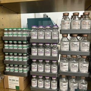 Op de afbeelding: Een verzameling doorzichtige glazen medische flesjes met gekleurde doppen, gerangschikt in grijze plastic trays op een plank. De etiketten tonen diverse medicijnnamen zoals Midazolam, Propofol, Ketamine, Lidocaïne, Ketorolac en Ondansetron.