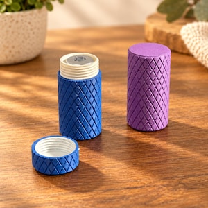Op de afbeelding: Twee cilindrische houders met een gestructureerd ruitpatroon aan de buitenkant. De ene houder is blauw met een bijpassend schroefdeksel en een open bovenkant die een witte binnenkant onthult. De tweede houder is paars met een gesloten deksel.