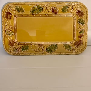May include: A rectangular ceramic serving platter in a glossy mustard yellow finish. The border features a raised, three-dimensional relief pattern of oak leaves and acorns in shades of green and brown, framed by a textured rope-like edge.