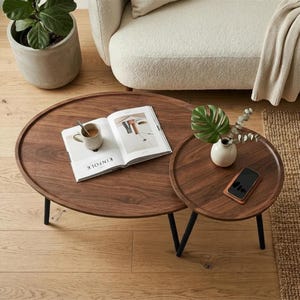 May include: A set of two nesting coffee tables made of dark brown wood with black metal legs. The larger oval table holds an open book titled Kinfolk and a ceramic mug. The smaller round table holds a white vase with green leaves and a smartphone.
