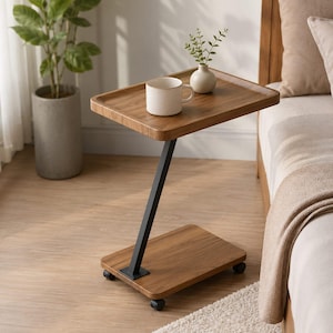 Peut inclure: Une table d'appoint en forme de C avec un plateau et une base au fini bois brun, reliés par un cadre en métal noir en forme de Z. La table est équipée de quatre petites roulettes noires. Un mug en céramique blanche et un petit vase blanc sont posés.