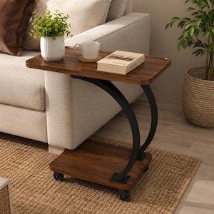 Peut inclure: Une table d'appoint en forme de C avec un plateau et une base au fini grain de bois foncé, soutenue par deux pieds métalliques noirs incurvés. La table dispose de quatre petites roulettes à la base. Un mug blanc, une plante et un livre sont posés.