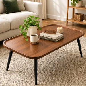 May include: A rectangular wooden coffee table with rounded corners and four tapered black legs. The table surface holds a potted green plant in a white ribbed ceramic pot, a white ceramic mug on a coaster, two stacked books, and a candle.