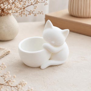 Peut inclure: Un bol en céramique blanc mat doté d'une figurine de chat sculptée sur le côté. Le chat est représenté en position de repos, les yeux fermés, enroulant ses pattes avant autour de l'extérieur du bol tandis que sa queue longe la base.