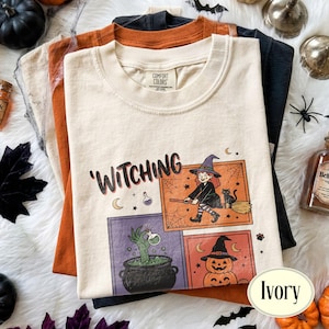 May include: An ivory t-shirt featuring the text 'WITCHING' and three illustrations: a witch on a broomstick with a cat, a green hand emerging from a cauldron, and stacked jack-o'-lanterns. The shirt has a Comfort Colors brand label at the neck.