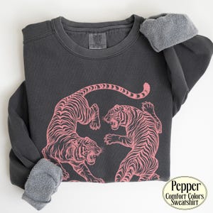 May include: A charcoal grey crewneck sweatshirt featuring a pink graphic of two tigers facing each other. The interior is lined with a grey fleece material. The neck label reads Comfort Colors, and a circular badge in the corner says Pepper Comfort Colors Sweatshirt.