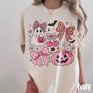 Könnte beinhalten: Ein cremefarbenes T-Shirt mit einem Grafikdruck eines Geistes mit einer Schleife im Leopardenmuster, einem rosa Kürbis mit Schleife, Kirschen, einem Spinnennetz, Fledermäusen, Herzen, Sternen und Blumen. Das Design enthält zudem eine große Leoparden-Schleife.