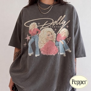 Può includere: Una t-shirt a maniche corte grigio antracite con una stampa grafica di Dolly Parton in tre pose diverse. Il design include il testo Dolly Parton, diverse forme a stella e il contorno di un cuore. La maglietta ha una vestibilità ampia e comoda.