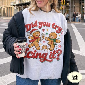 May include: A light gray crewneck sweatshirt featuring a graphic of two gingerbread cookies with the text Did you try icing it? One cookie wears a Santa hat and scarf, while the other appears broken. The design includes red and green accents.