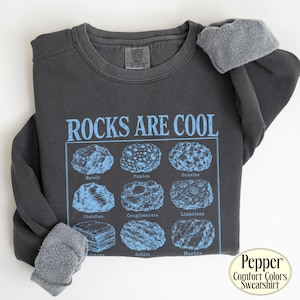 Może przedstawiać: Ciemnoszara bluza z okrągłym dekoltem z jasnoniebieskim nadrukiem graficznym z napisem ROCKS ARE COOL nad siatką dziewięciu okazów geologicznych oznaczonych jako Basalt, Pumice, Granite, Obsidian, Conglomerate, Limestone, Sandstone, Schist i Marble.