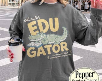 Può includere: Una felpa girocollo grigio antracite con la grafica di un alligatore verde chiaro che indossa occhiali e legge un libro aperto. Il testo recita "educator EDU GATOR" in grandi lettere gialle e "swamp-tested. classroom approved." in bianco.