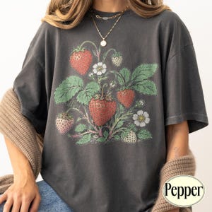 May include: A charcoal gray short-sleeve t-shirt featuring a botanical illustration of red strawberries, green leaves, and white flowers centered on the chest. The graphic displays multiple strawberries in various stages of ripeness.
