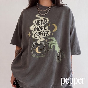 Può includere: Una t-shirt grigio antracite a maniche corte con una stampa grafica di una mano verde che regge una tazza fumante. Il testo "NEED MORE COFFEE" è stampato in bianco sopra la tazza, accompagnato da illustrazioni dorate di lune e stelle.