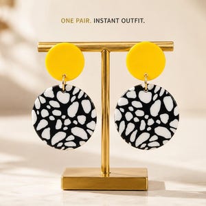 May include: A pair of dangle earrings displayed on a gold-toned T-bar stand. Each earring features a solid yellow circular top piece connected to a larger circular bottom piece with a black and white abstract spotted pattern. Text reads: ONE PAIR. INSTANT OUTFIT.