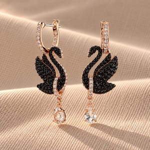 May include: A pair of rose gold-tone earrings featuring a swan motif encrusted with small black crystals. The swan bodies are accented with clear crystals along the neck and hoop, and each earring includes a dangling clear pear-shaped crystal drop.